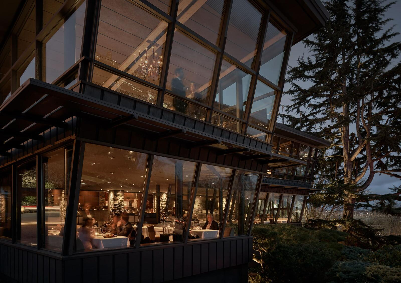 Canlis restaurant at dusk, glass window bays glowing warm above a hillside of evergreens